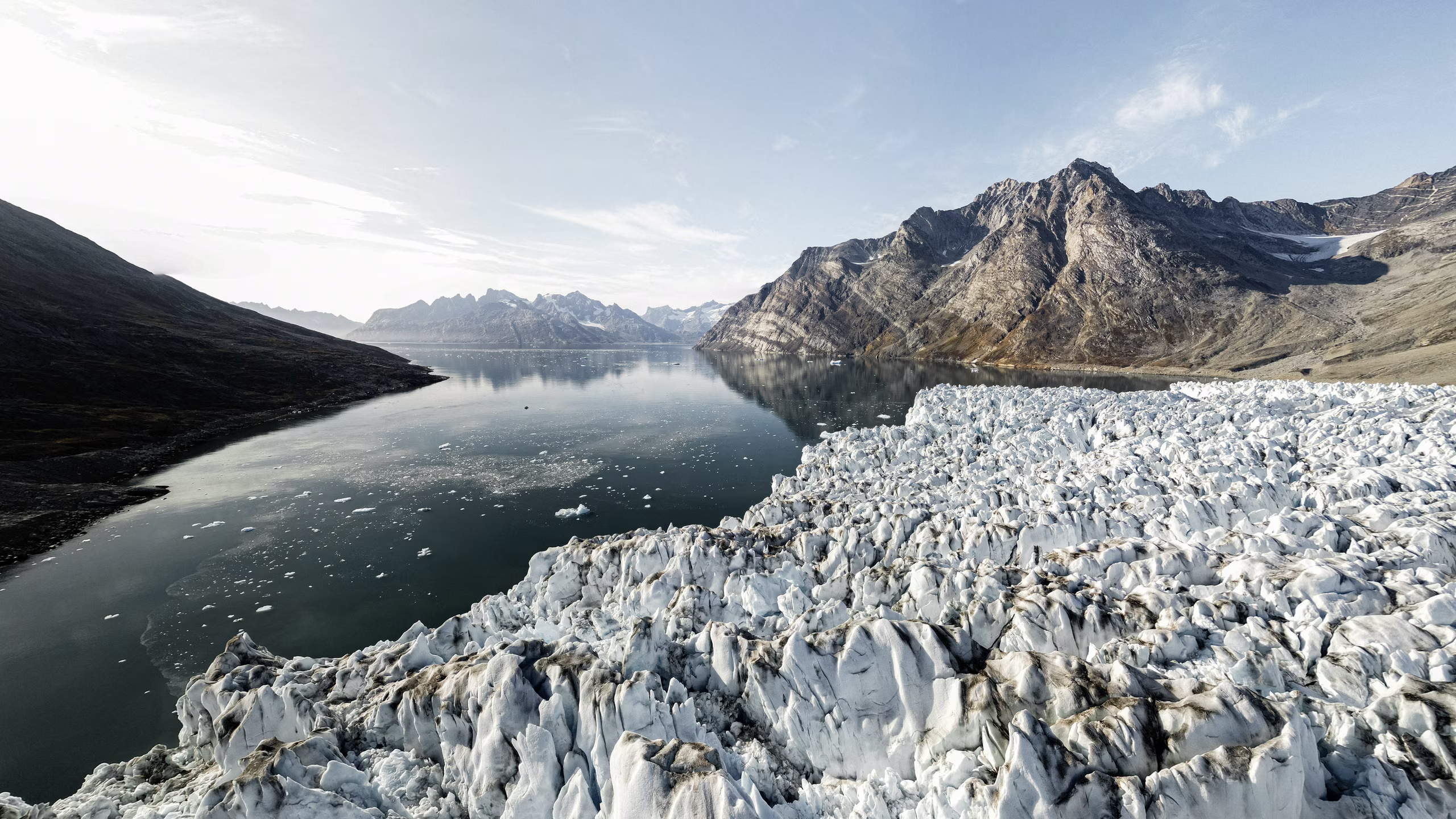 Glacier ice flows into calm fjord water surrounded by rugged mountains under a clear sky.