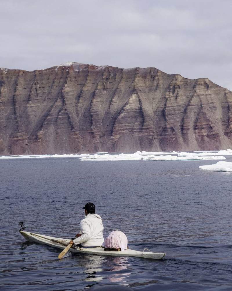 Person paddling a kayak on calm water with icebergs and a rocky cliff in the background.