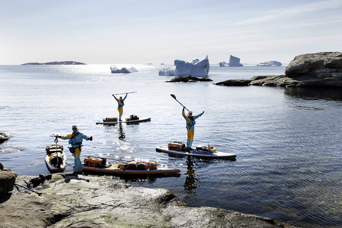 Three people in wetsuits on paddleboards by rocky shore with icebergs in calm ocean water.