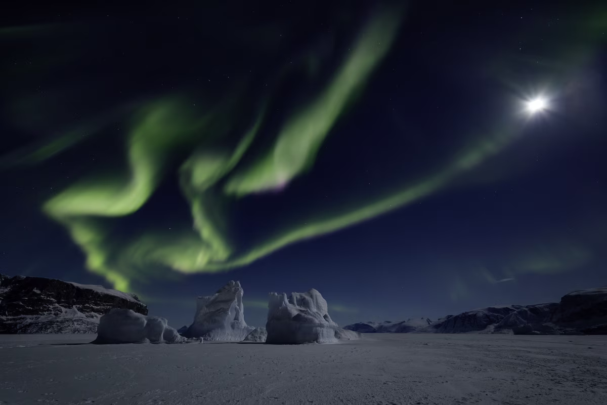 Green northern lights swirl over snowy icebergs and a dark mountainous landscape at night.