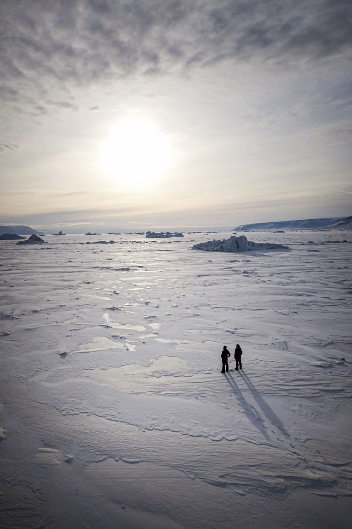 Two people stand on a vast frozen landscape under a cloudy sky with the sun low on the horizon.