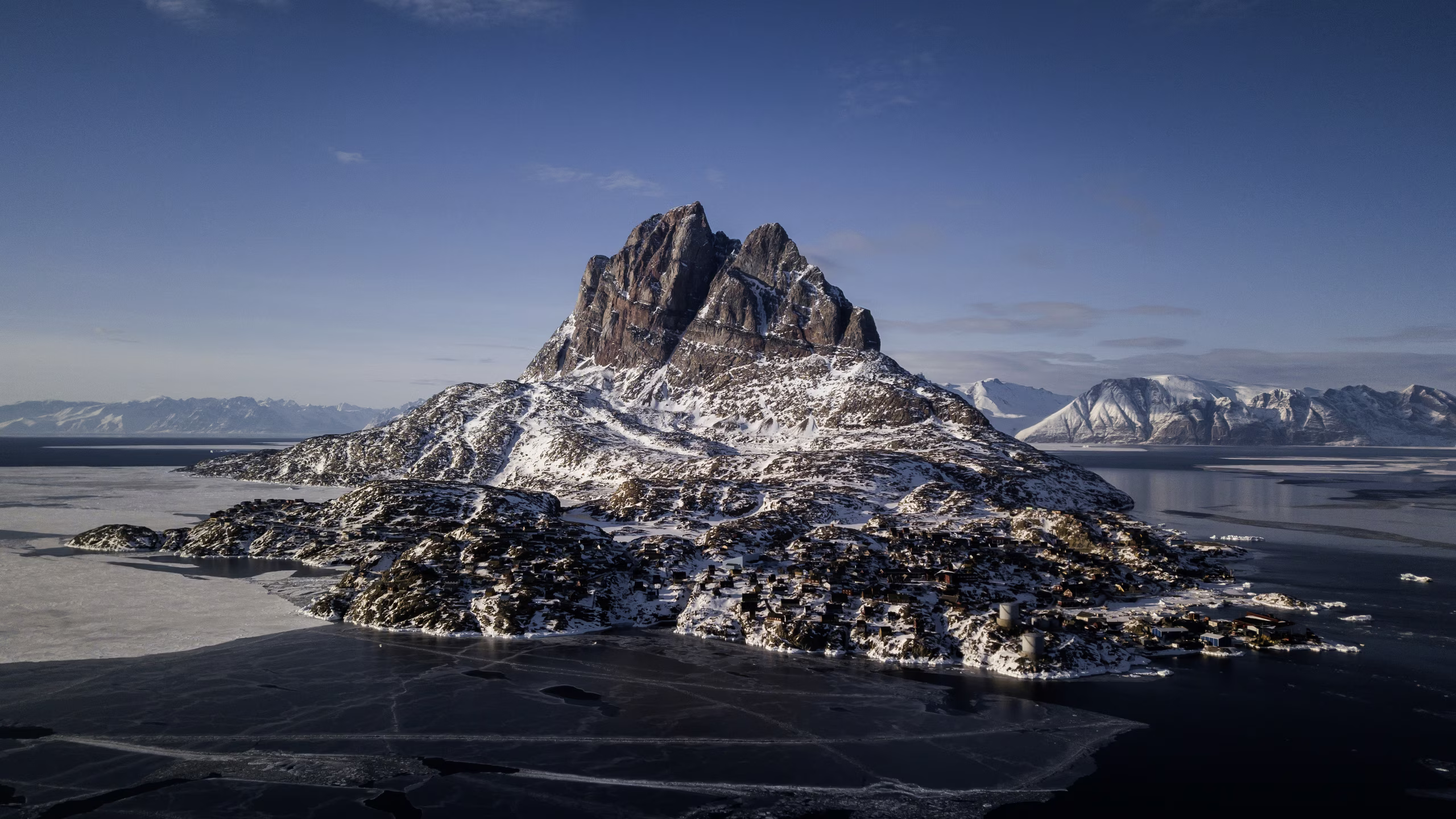 Snow-covered island with rocky mountain peak surrounded by icy sea under a clear blue sky.