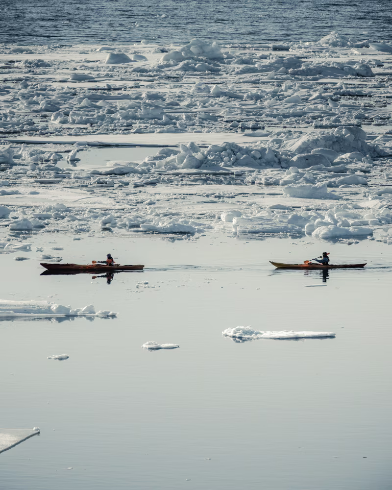 Two kayakers paddling on calm icy water surrounded by floating ice chunks and snow.