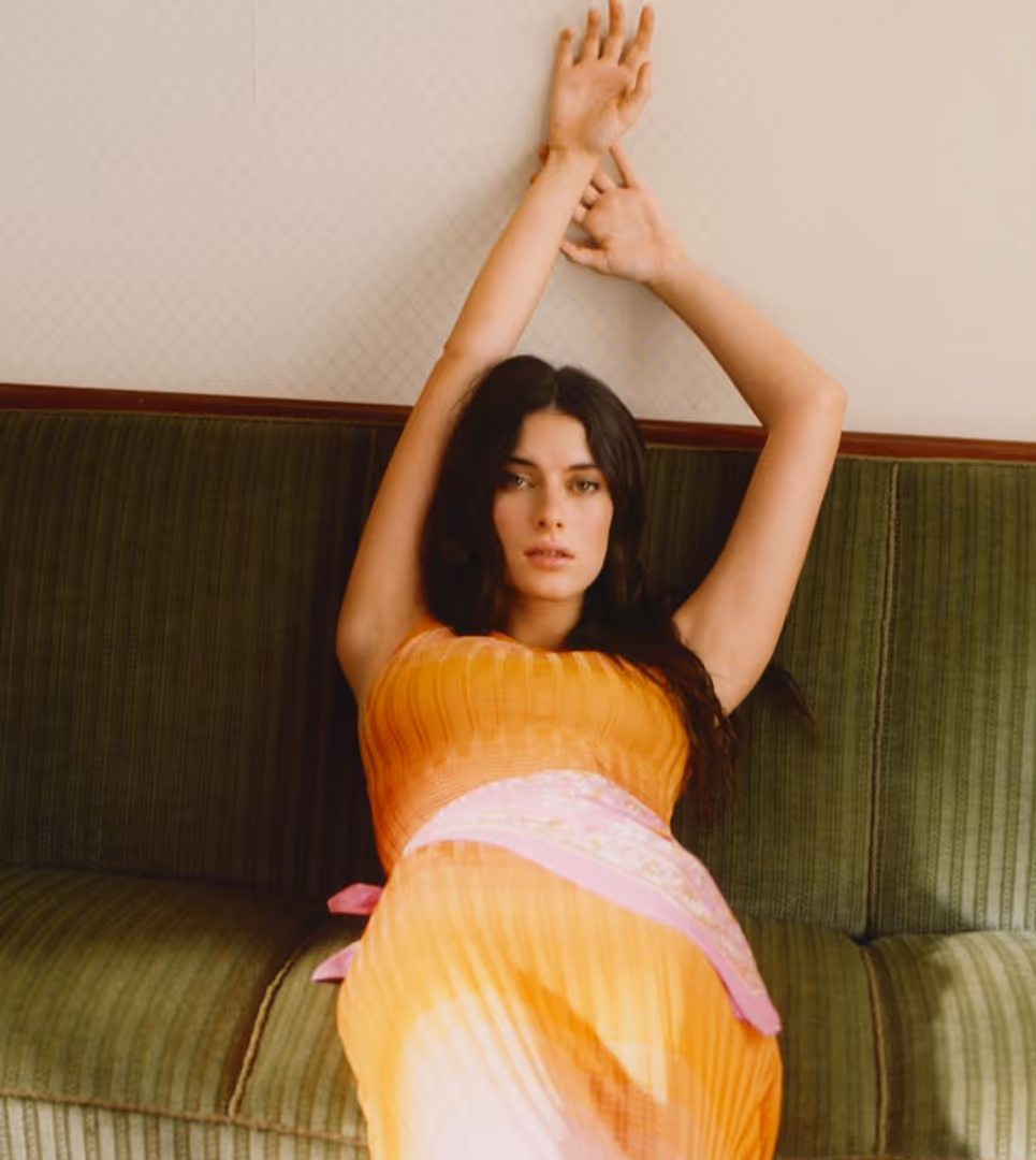 Woman in orange dress with a pink sash reclining on a green sofa with arms raised above her head.