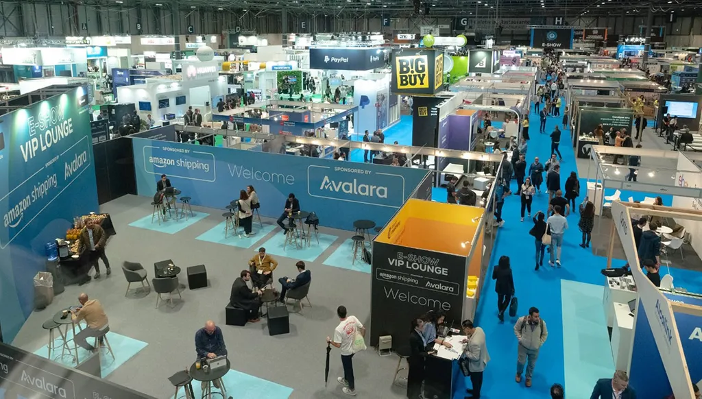 A large convention hall with booths and people walking on blue carpet at a technology expo featuring Amazon Shipping and Avalara VIP Lounge.