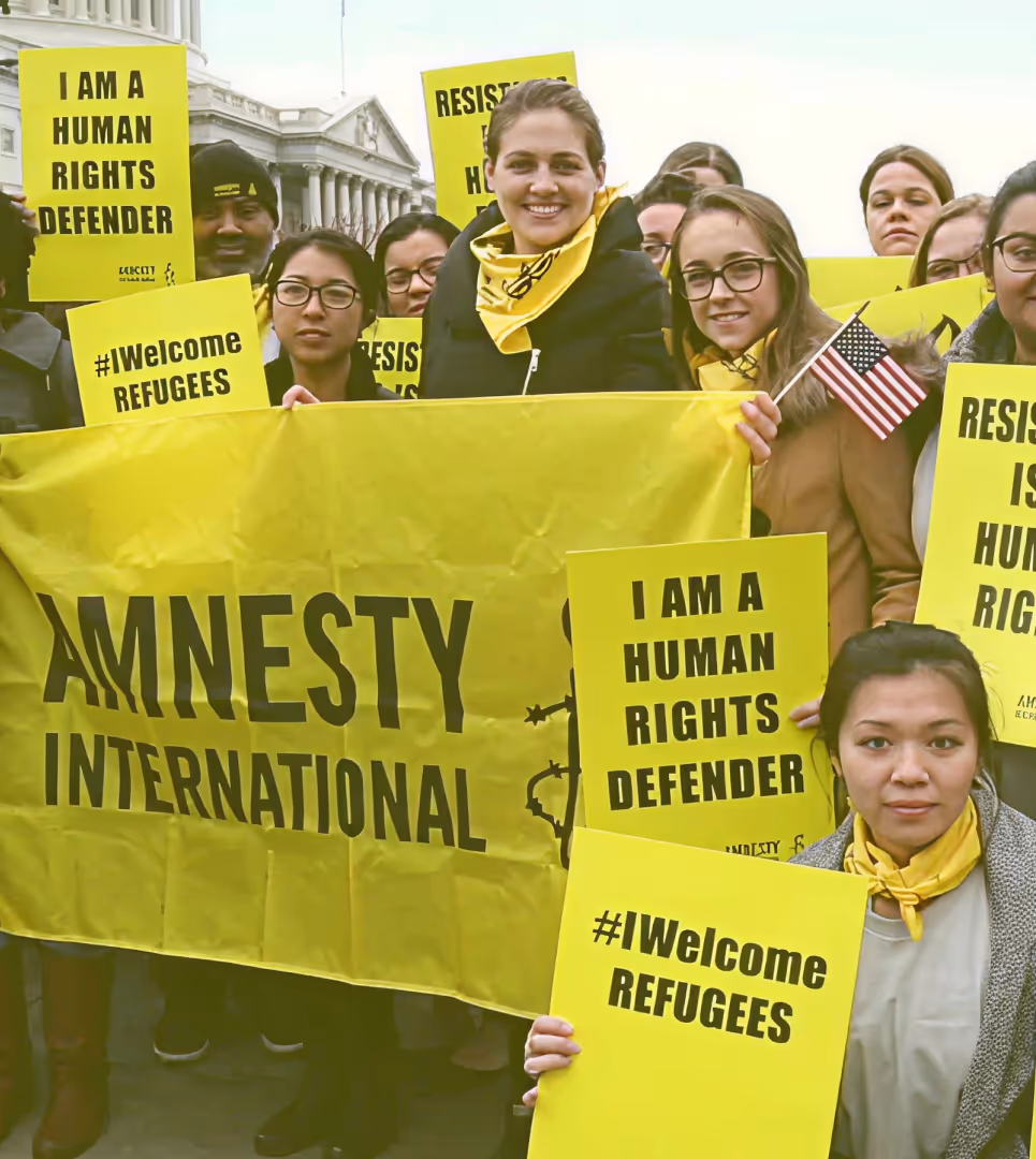 Group of diverse activists holding yellow Amnesty International banners and signs saying 'I am a human rights defender' and '#Welcome Refugees' in front of a government building.