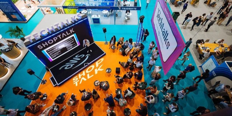 Overhead view of a tech conference session with a speaker on stage presenting to an audience seated on orange flooring, surrounded by booths and attendees walking around.