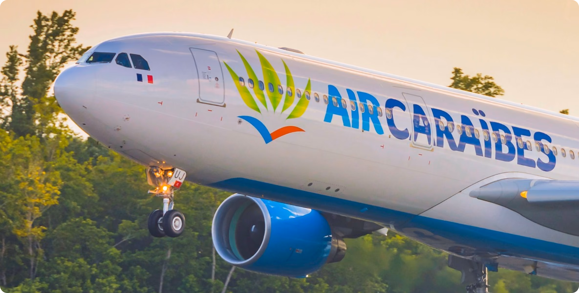 Air Caraïbes commercial airplane landing or taking off with landing gear down against a background of green trees and a light sky at sunset or sunrise.