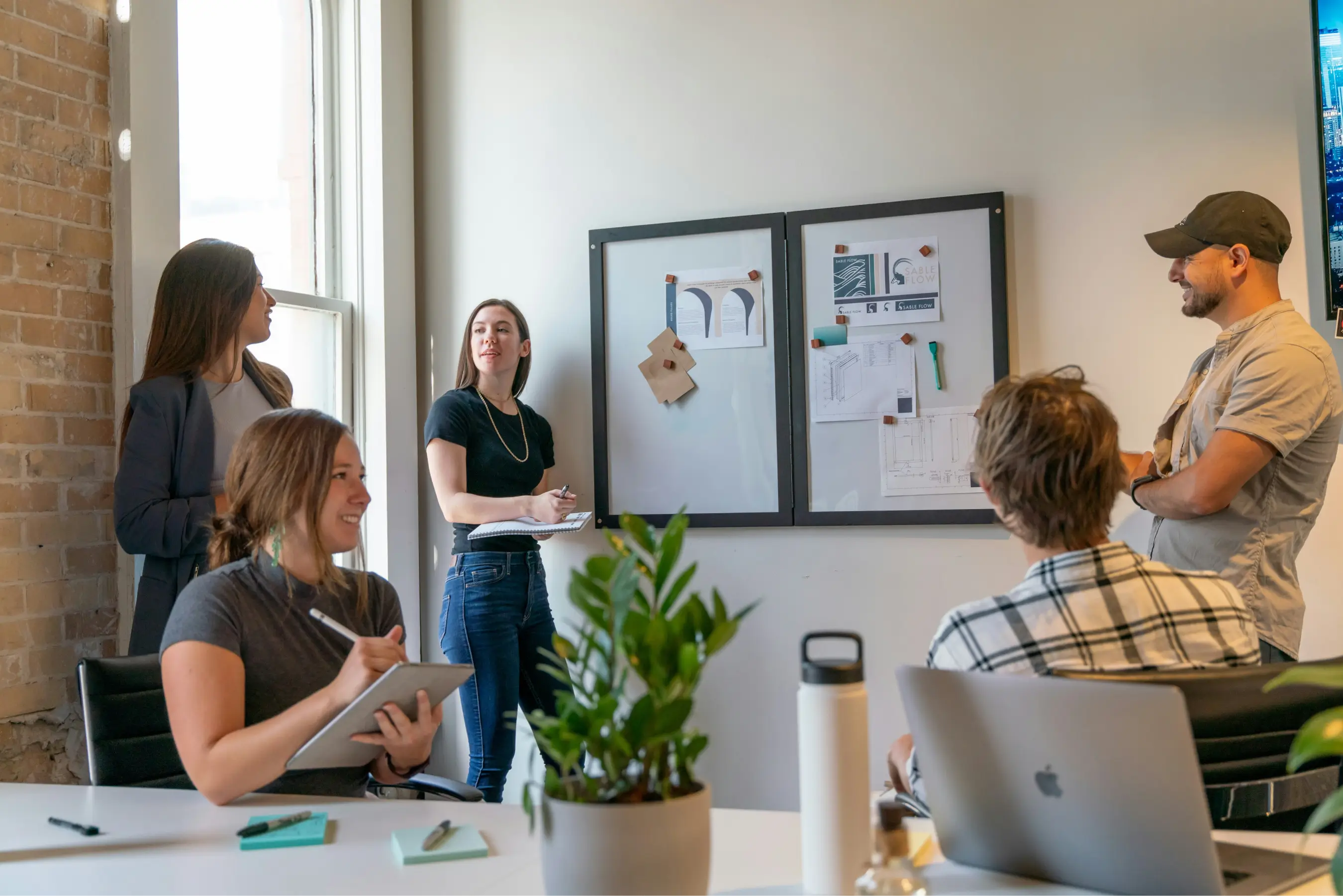 Five people engaged in a meeting in an office, with notes on a whiteboard and laptops nearby.