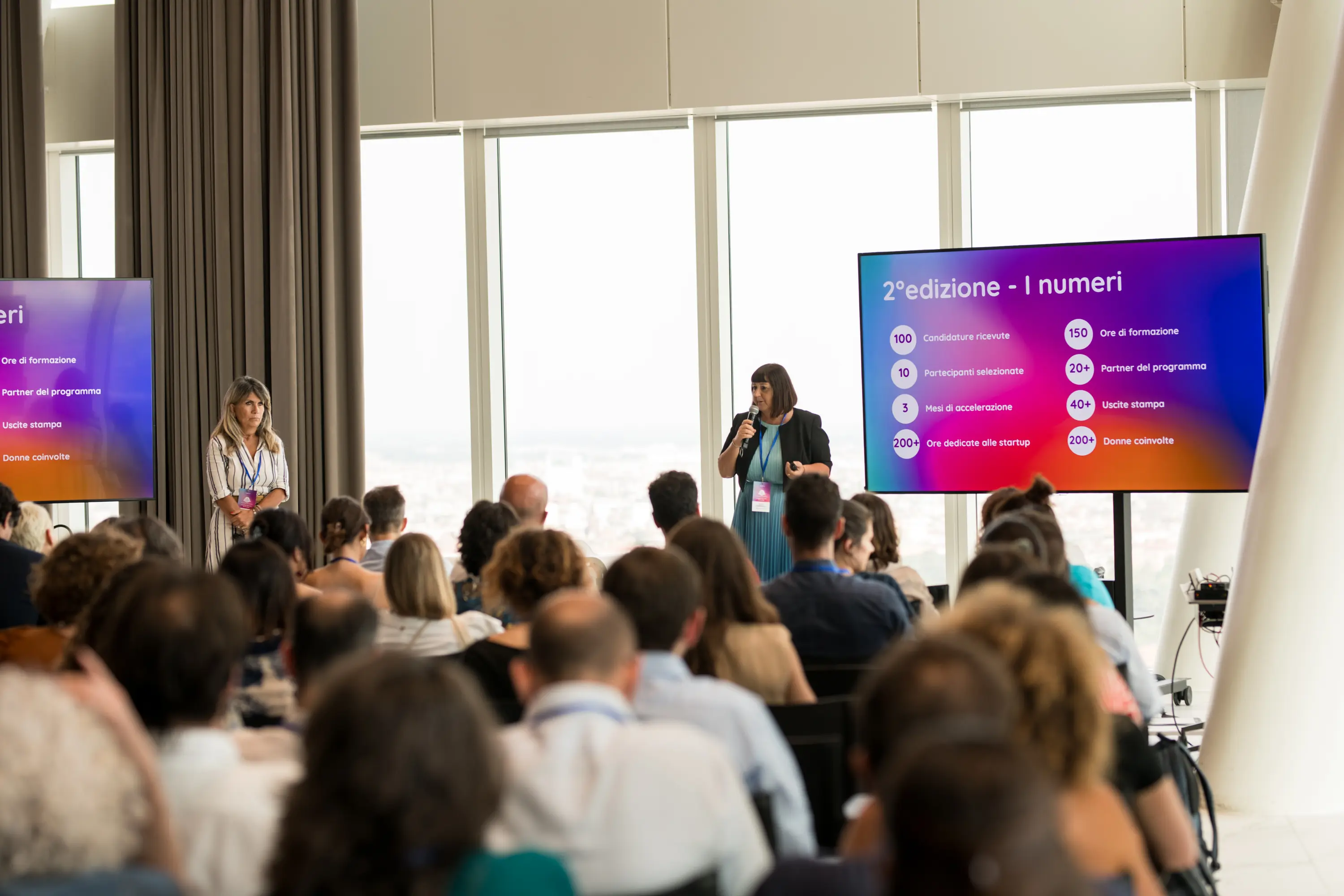 Evento Woman on Action: Donna che parla con un microfono a una conferenza davanti a un pubblico, con un grande schermo che mostra statistiche sulla seconda edizione di un programma in italiano.