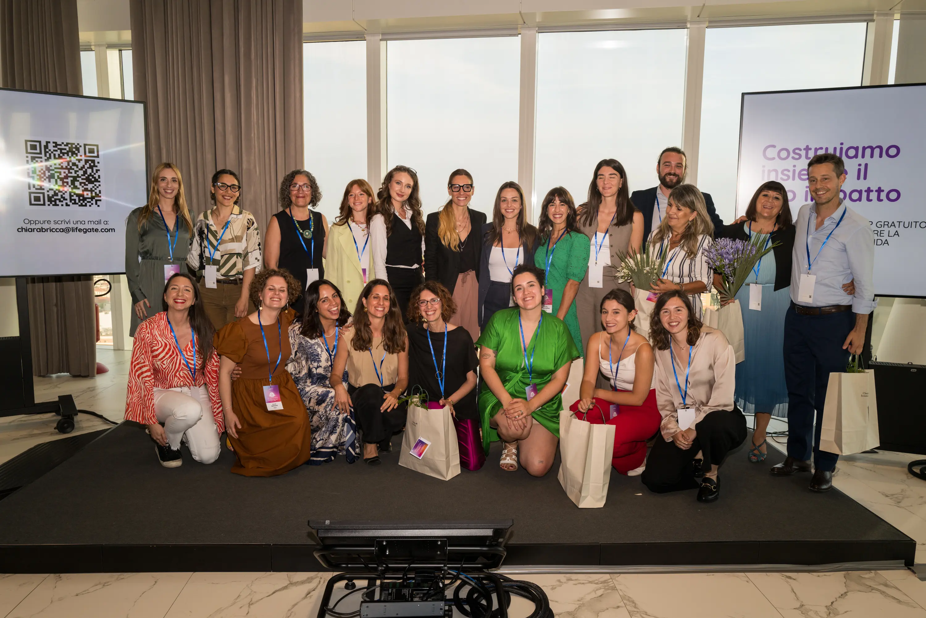 Evento Woman on Action: Gruppo di 20 persone che posano su un palco con badge dell’evento, alcune con fiori e borse regalo, davanti a grandi finestre e due schermi.