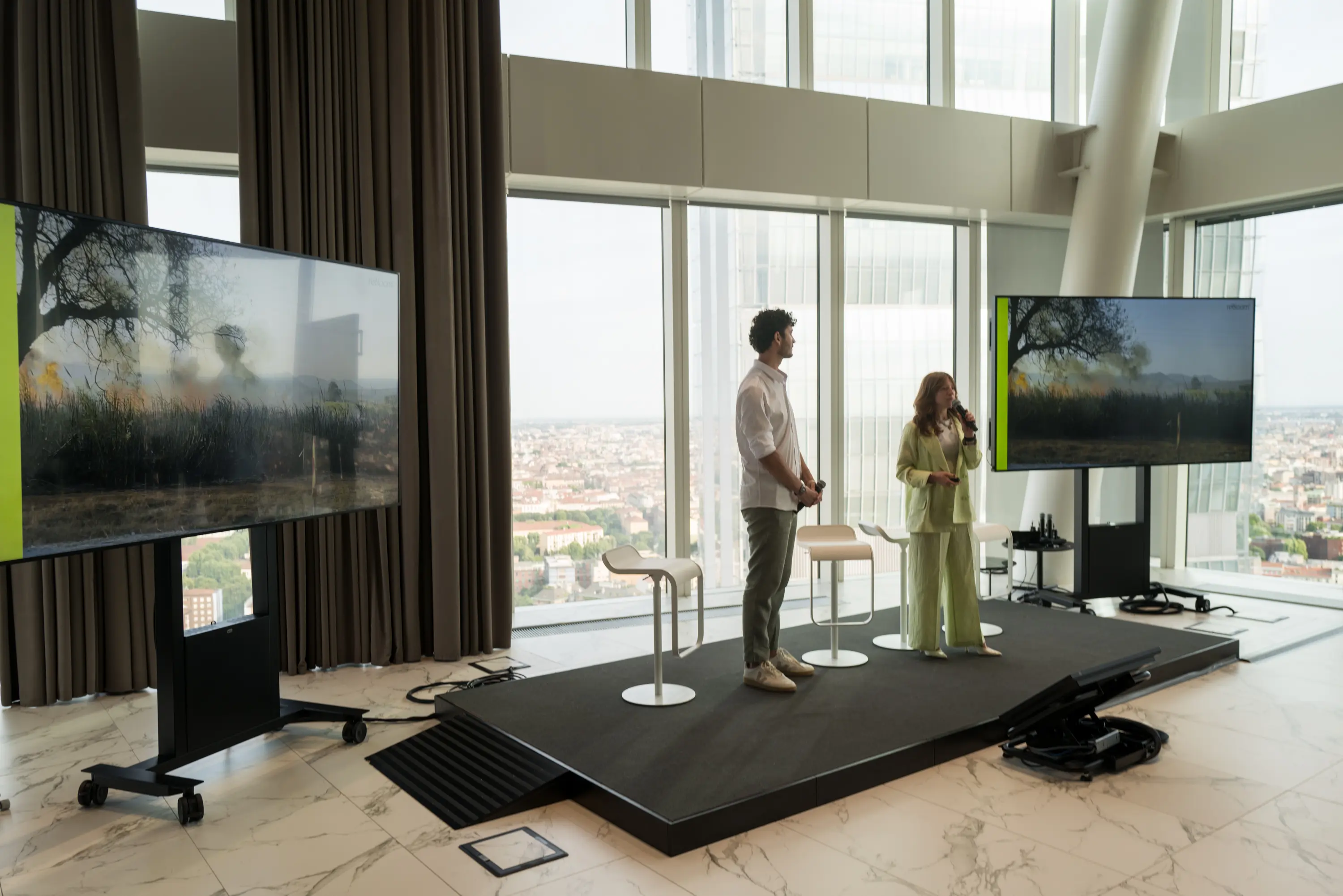 Evento Woman on Action: Uomo e donna su un palco in una stanza moderna con grandi finestre, due schermi che mostrano un paesaggio con alberi e fuoco, e sgabelli bianchi.