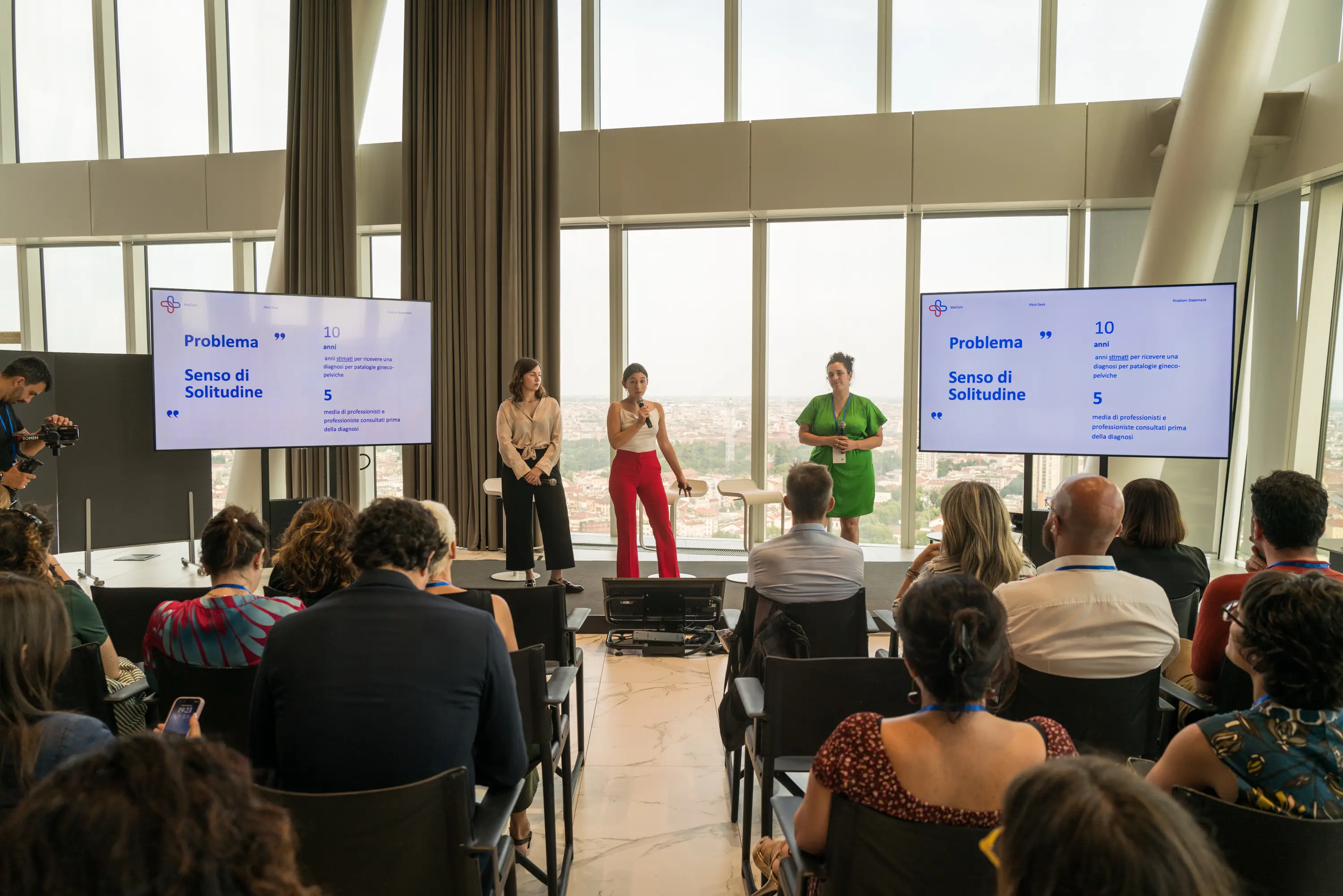 Evento Woman on Action: Tre donne presentano sul palco davanti a un pubblico con due schermi che mostrano una slide intitolata 'Problema Senso di Solitudine' in una stanza luminosa con grandi finestre con vista sulla città.