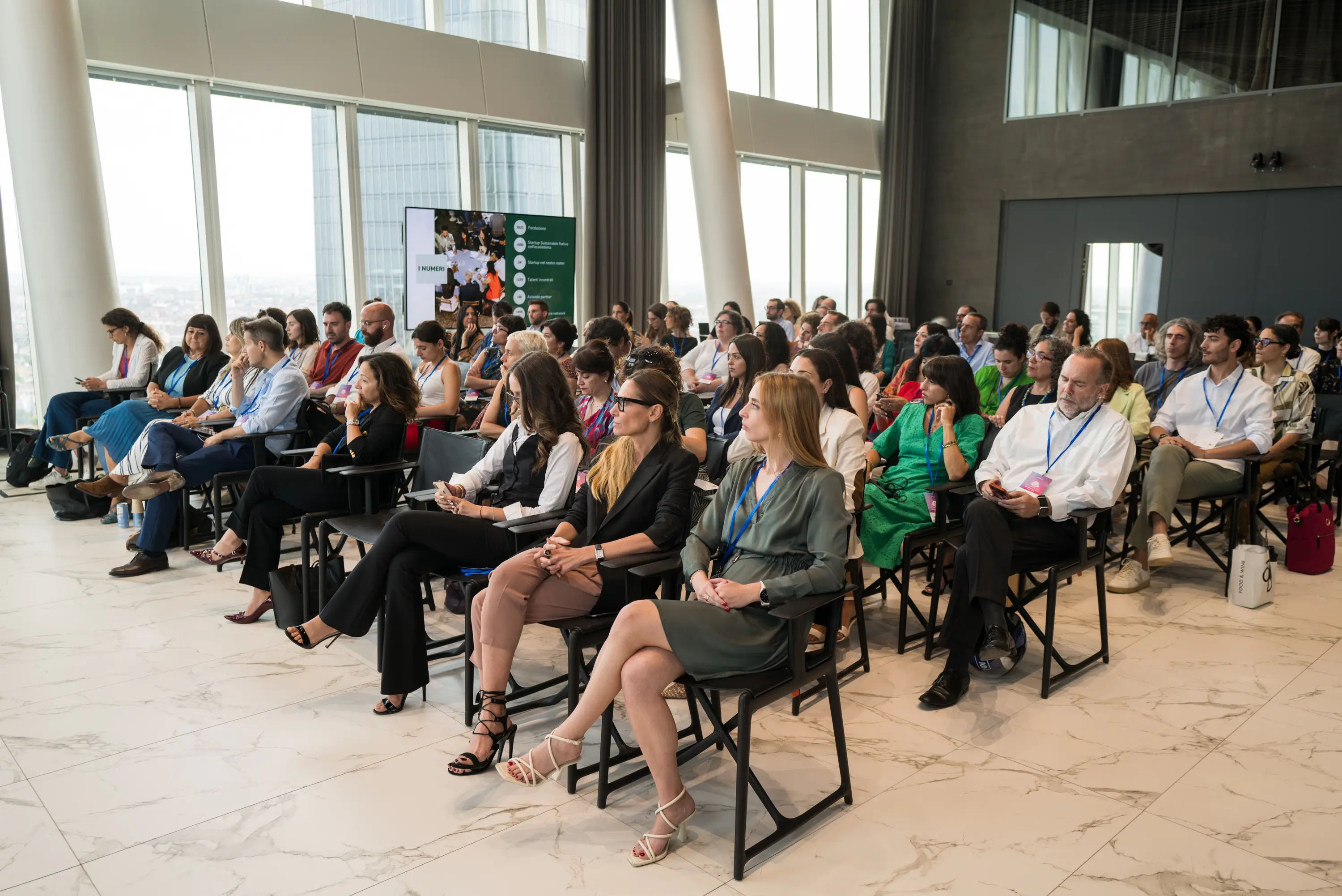 Evento Woman on Action: Gruppo diversificato di persone sedute attentamente in una moderna sala conferenze con grandi finestre e uno schermo per la presentazione.