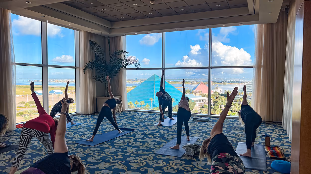 A yoga class with arms raised in the Floral Ballroom at Moody Gardens, with the pyramids and Galveston Bay visible through the windows