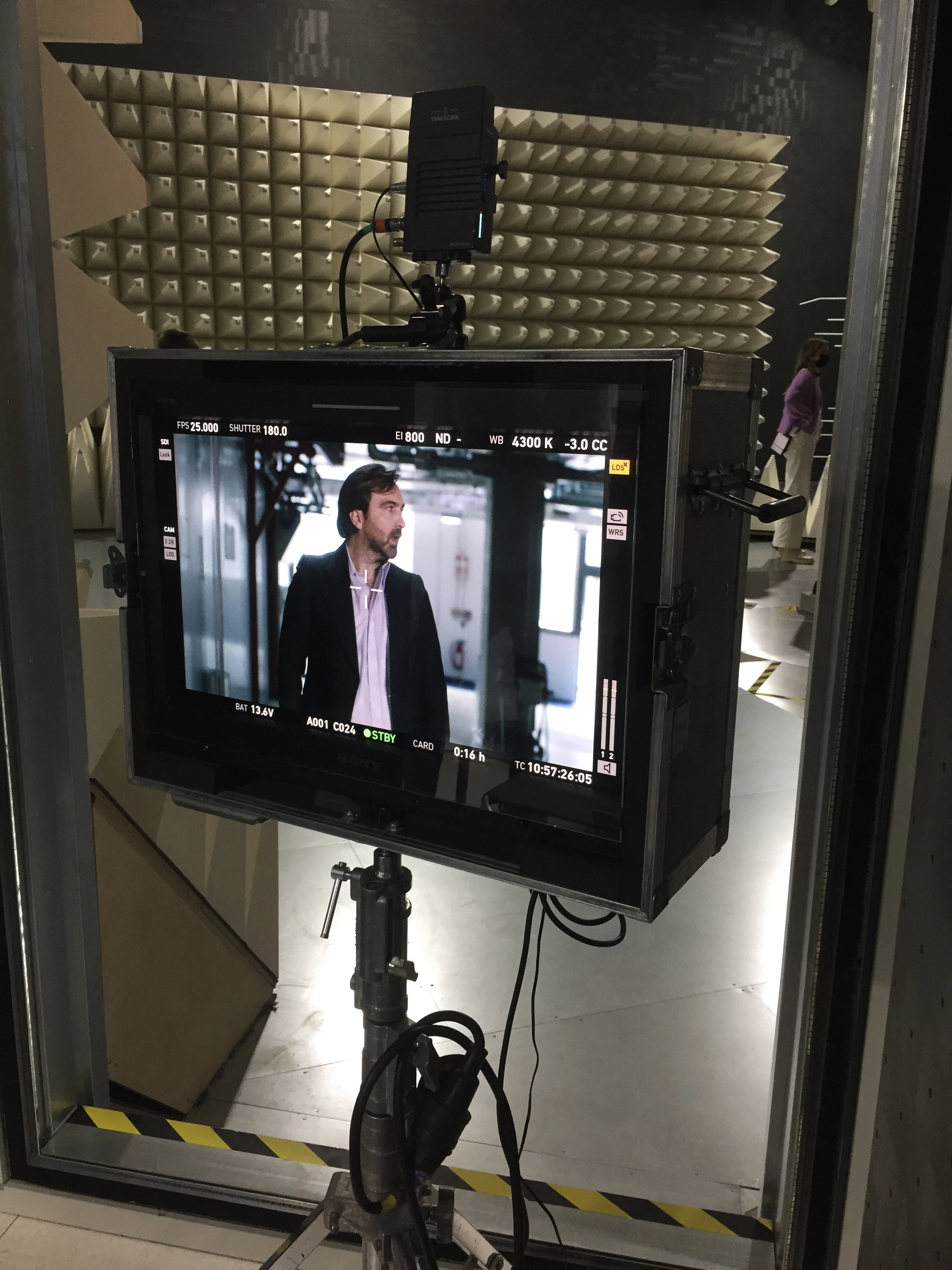 Monitor zeigt eine Filmaufnahme eines Mannes mit schwarzem Jackett und weißem Hemd vor einem unscharfen Hintergrund in einem Studio mit Schallschutzpaneelen.