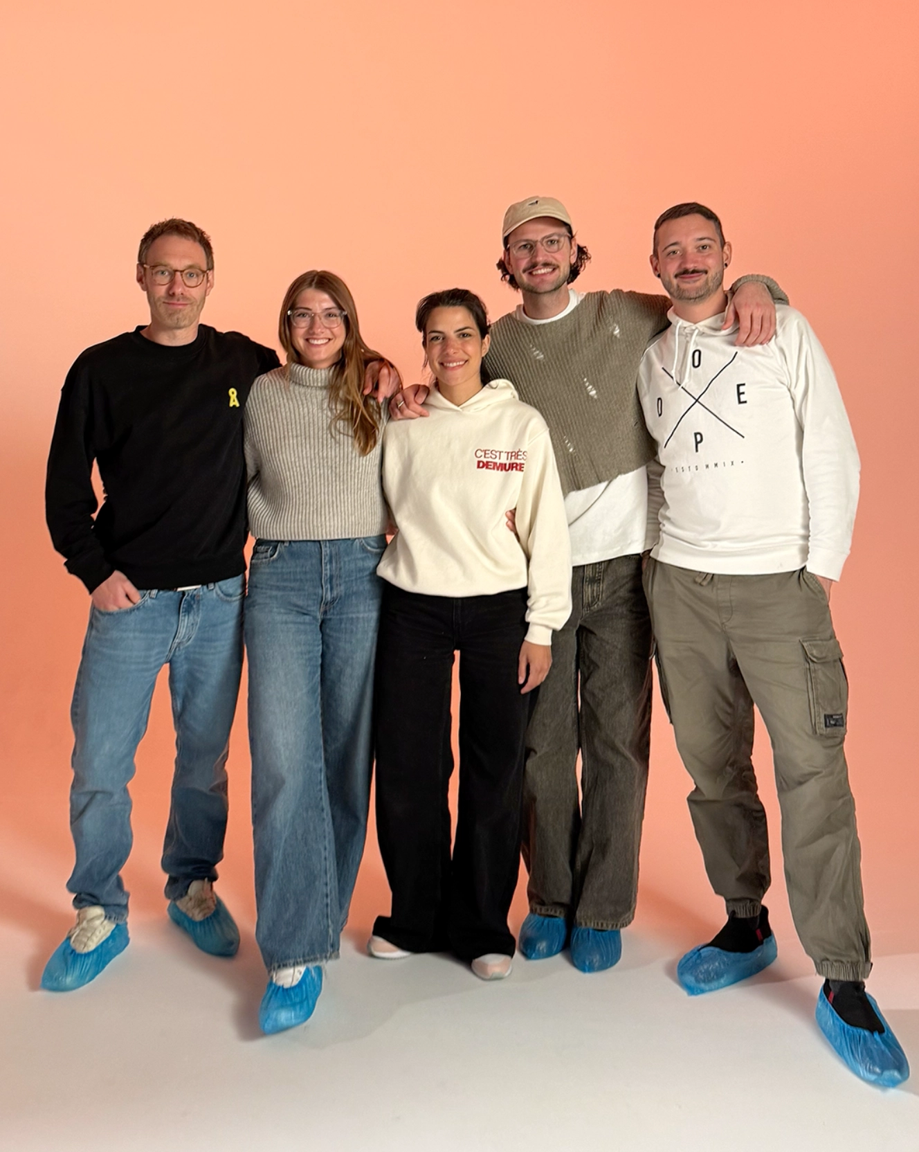 Fünf Personen stehen eng nebeneinander vor einem orangen Hintergrund, alle tragen blaue Überschuhe.