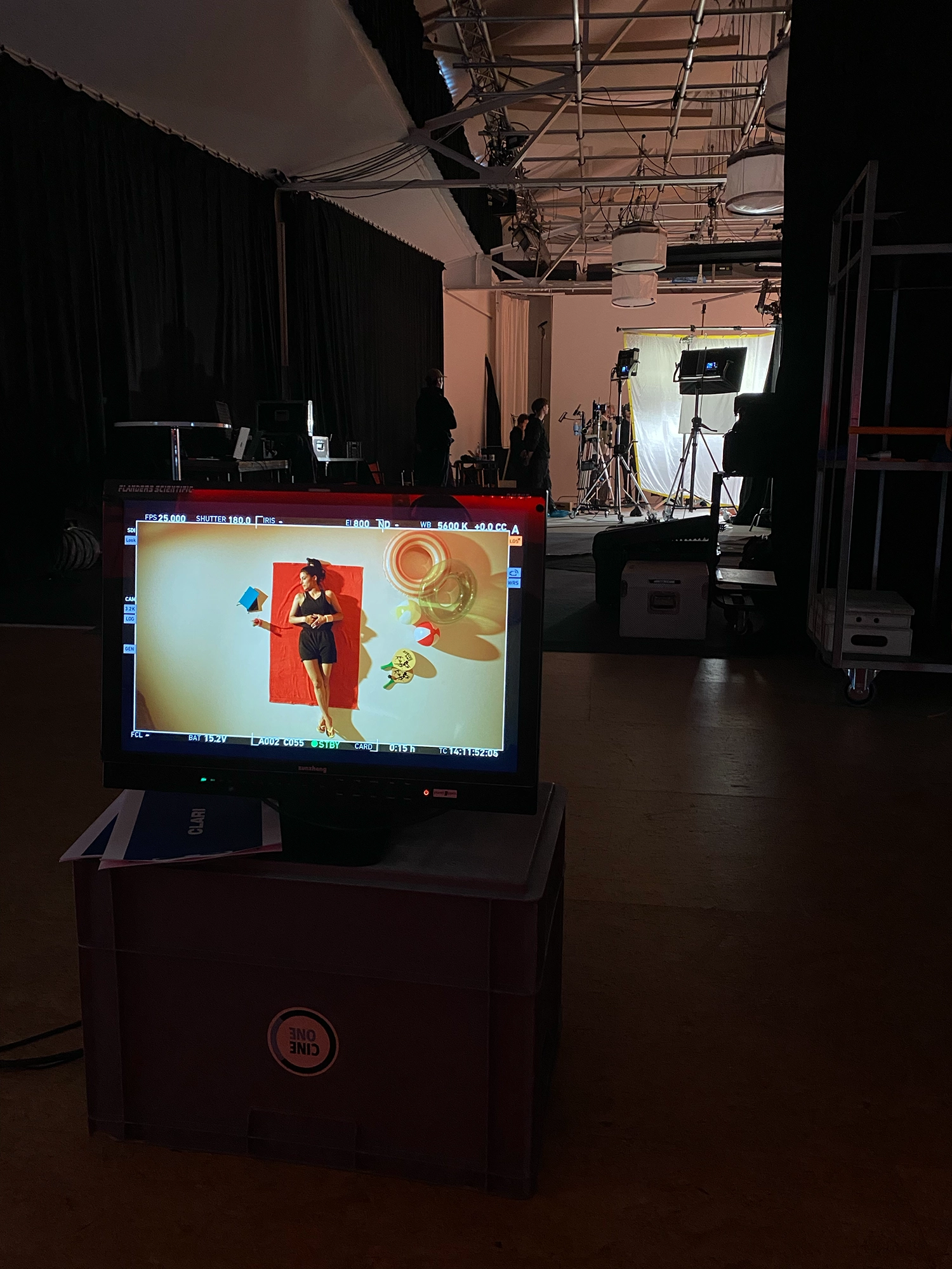 Studioaufnahme mit Bildschirm, der eine Frau zeigt, die auf einem roten Handtuch liegt, umgeben von Strandspielzeug, während Fotografen im Hintergrund arbeiten.