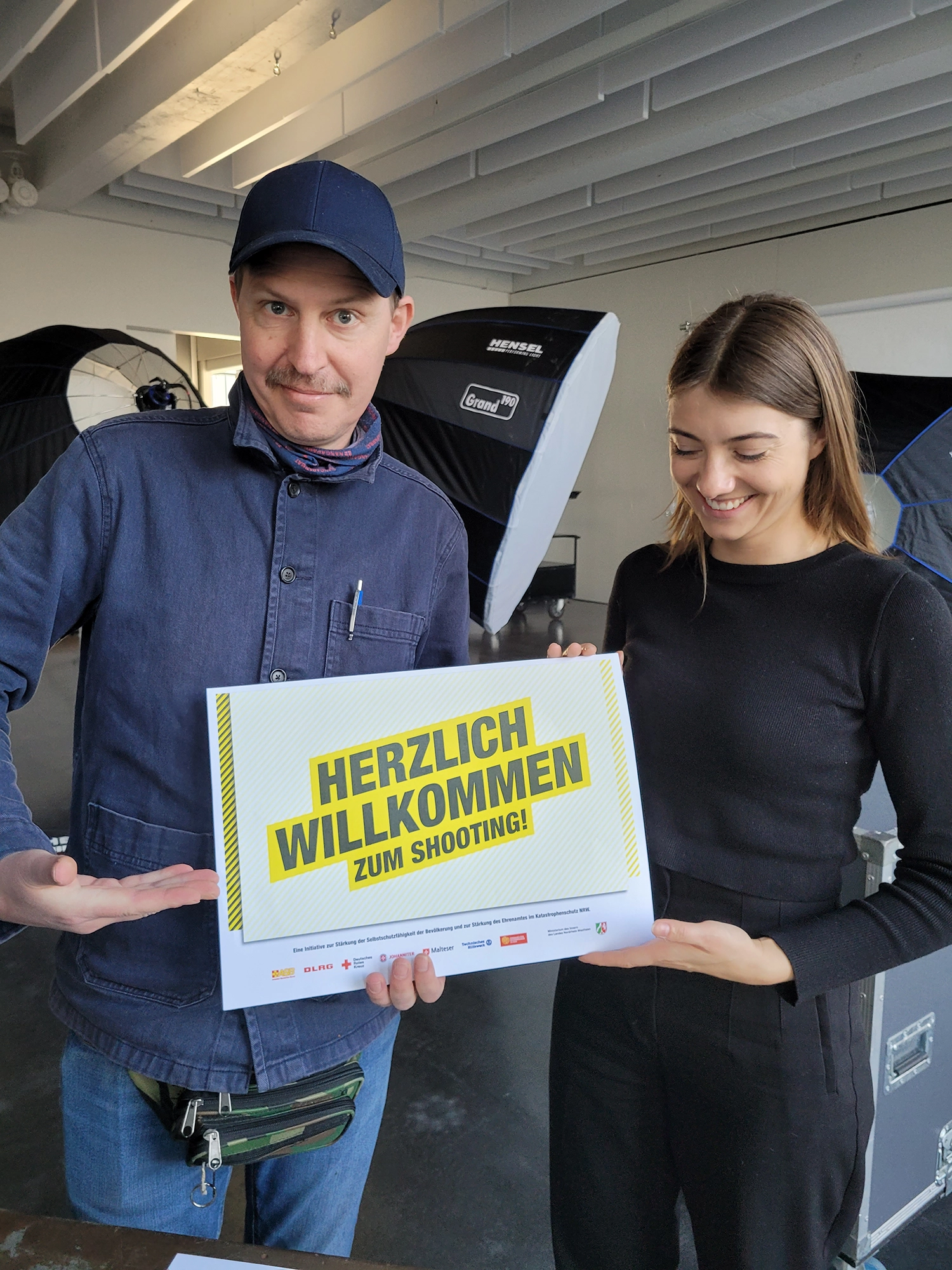 Ein Mann und eine Frau halten ein Schild mit der Aufschrift ‚Herzlich Willkommen zum Shooting!‘ in einem Fotostudio.
