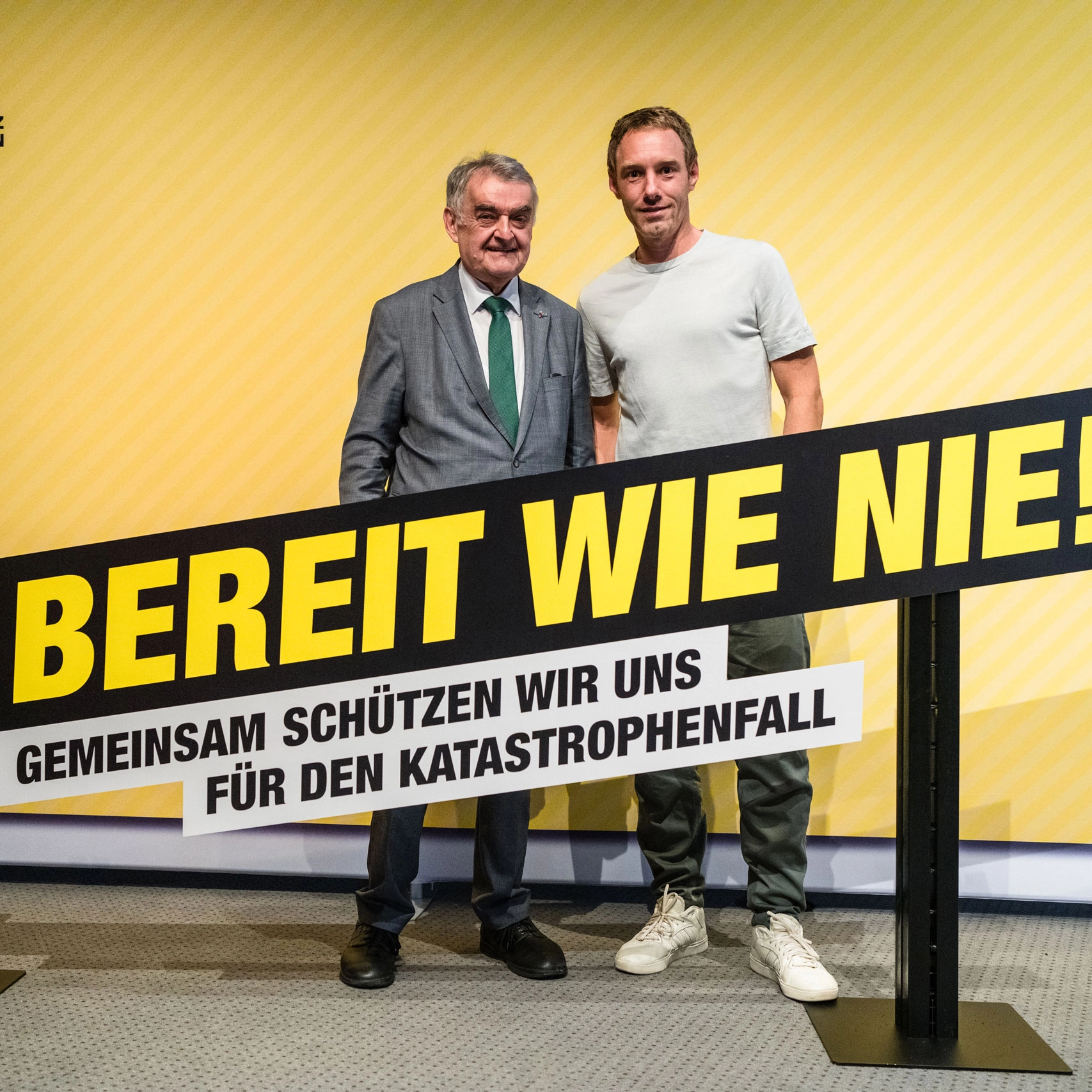 Zwei Männer, einer im Anzug und einer in weißem T-Shirt, stehen vor einem gelben Hintergrund mit dem Schild BEREIT WIE NIE! GEMEINSAM SCHÜTZEN WIR UNS FÜR DEN KATASTROPHENFALL.