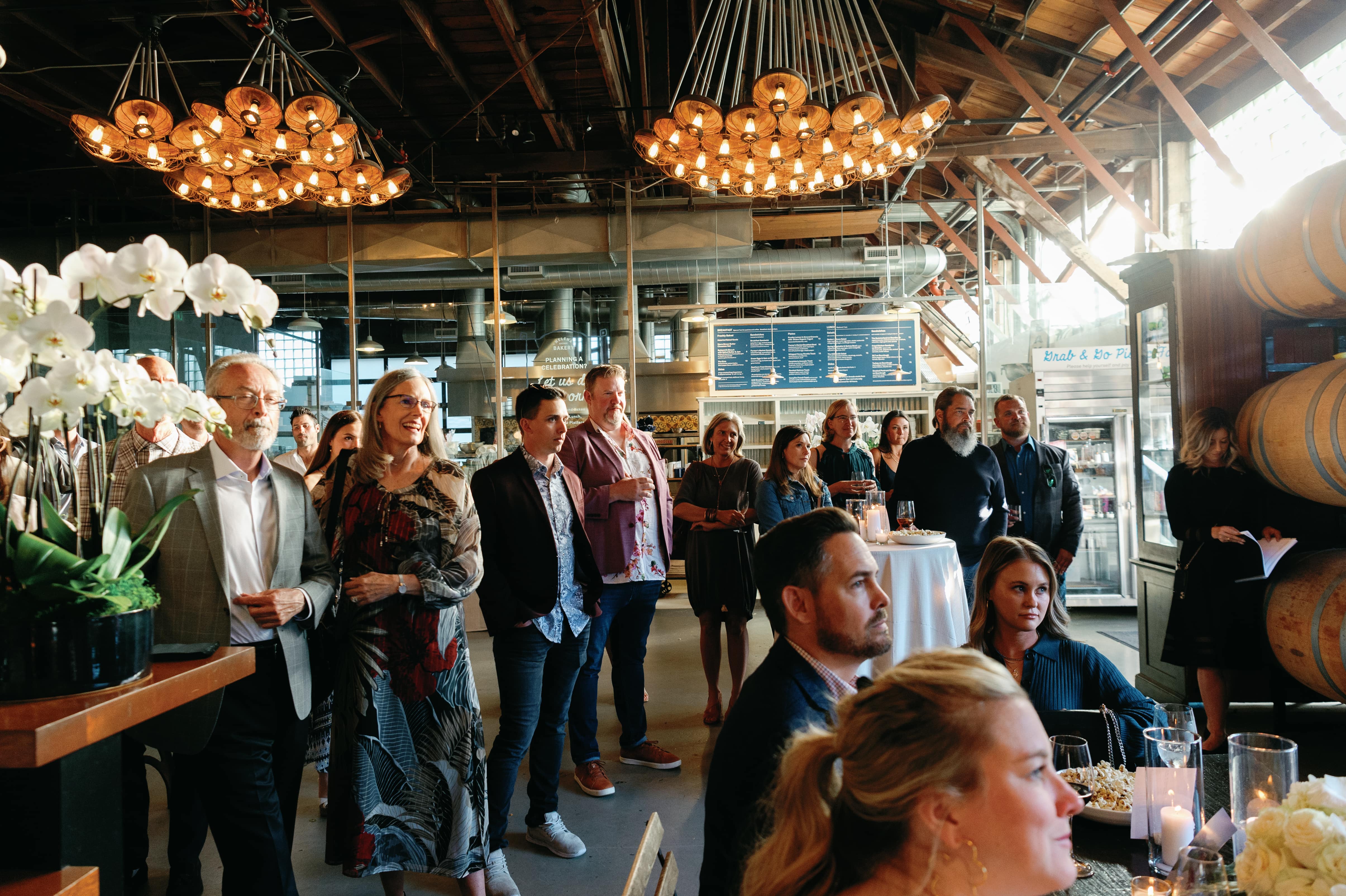 Well-dressed crowd with drinks in an industrial venue lined with wine barrels