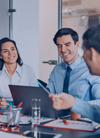 A group of three business individuals engaged in discussion at a table with a laptop open in front of them.