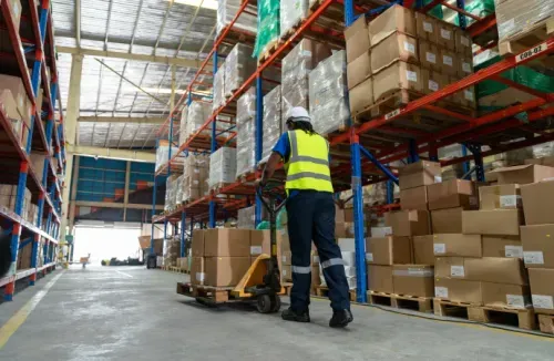Warehouse worker in safety vest and helmet pushing a pallet jack with boxes amidst tall shelves of packed pallets.