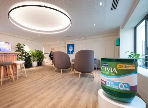 Modern office lounge with round ceiling light, chairs, plants, and a large Activia yogurt cup model.