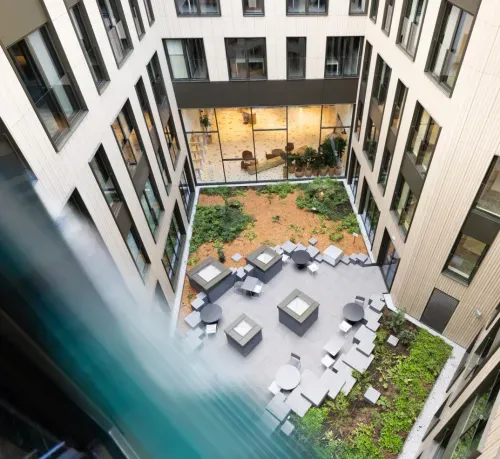 Modern building courtyard with garden beds, patio tables, chairs, and large windows overlooking inside.