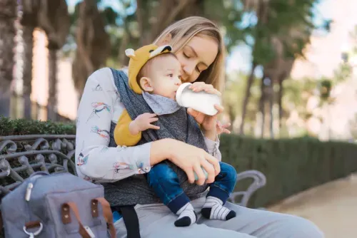 Mother sitting on a bench outdoors feeding baby from a bottle while holding the child in a carrier.