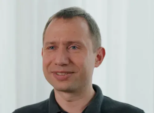 Smiling man with short brown hair wearing a dark polo shirt against a light background.