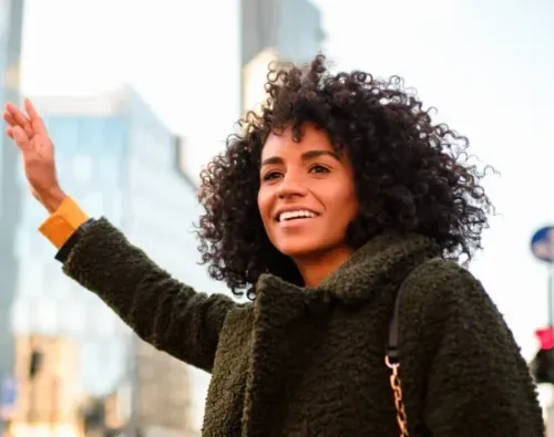 Smiling woman with curly hair raising her arm outdoors in a city to hail a ride.