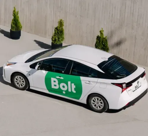 White sedan with green Bolt logo on door parked outside next to concrete wall with three potted bushes.
