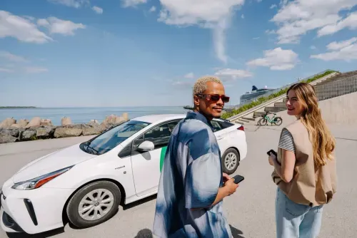 Two people holding phones next to a white hybrid car by the water on a sunny day.