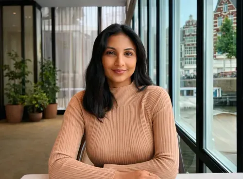 Woman with dark hair wearing a tan ribbed sweater sitting indoors near large windows with plants nearby.