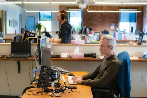 Man with gray hair types on a keyboard at a workstation with dual monitors in an open office.