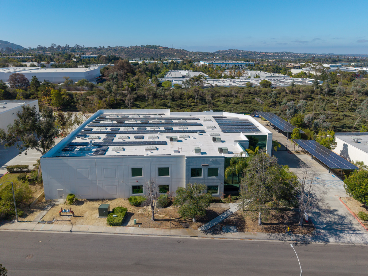 Aerial view of a solar-paneled commercial building with additional ground-mounted solar panel rows beside it, set among an industrial park with hills, trees, and mountains stretching to the horizon.