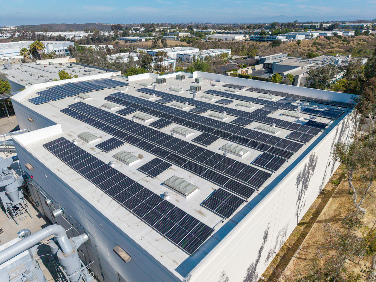Close-up aerial view of a flat commercial rooftop densely covered with solar panels arranged around HVAC units and skylights, with an industrial park visible below.
