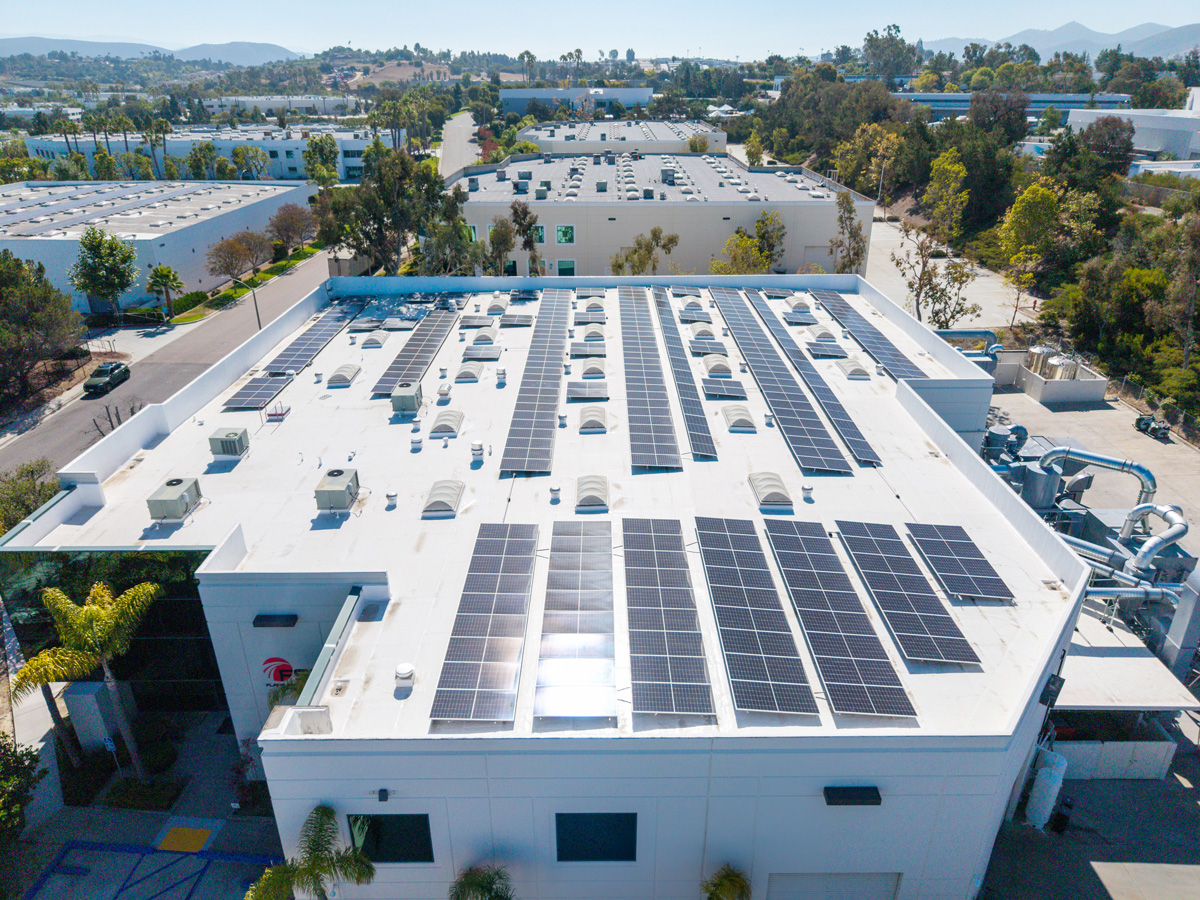 Aerial view of a commercial building with rooftop and ground-mounted solar panels, located at a street corner in a business park