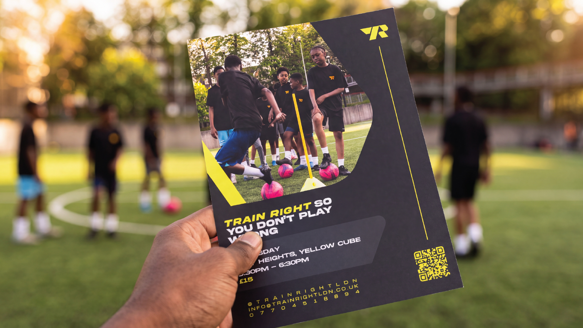 Hand holding a flyer for youth soccer training with kids practicing on a field in the background at sunset.