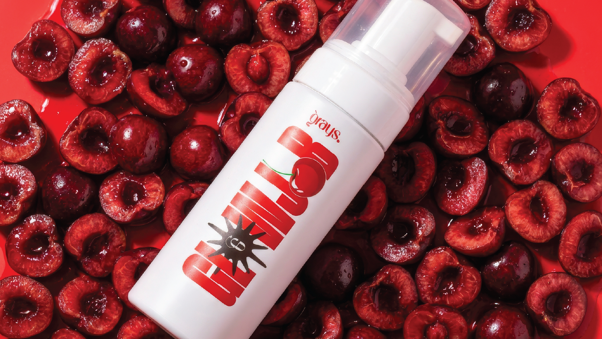 White pump bottle labeled 'Cherry Bomb' with a cherry graphic, lying on a bed of halved and whole fresh cherries on a red surface.