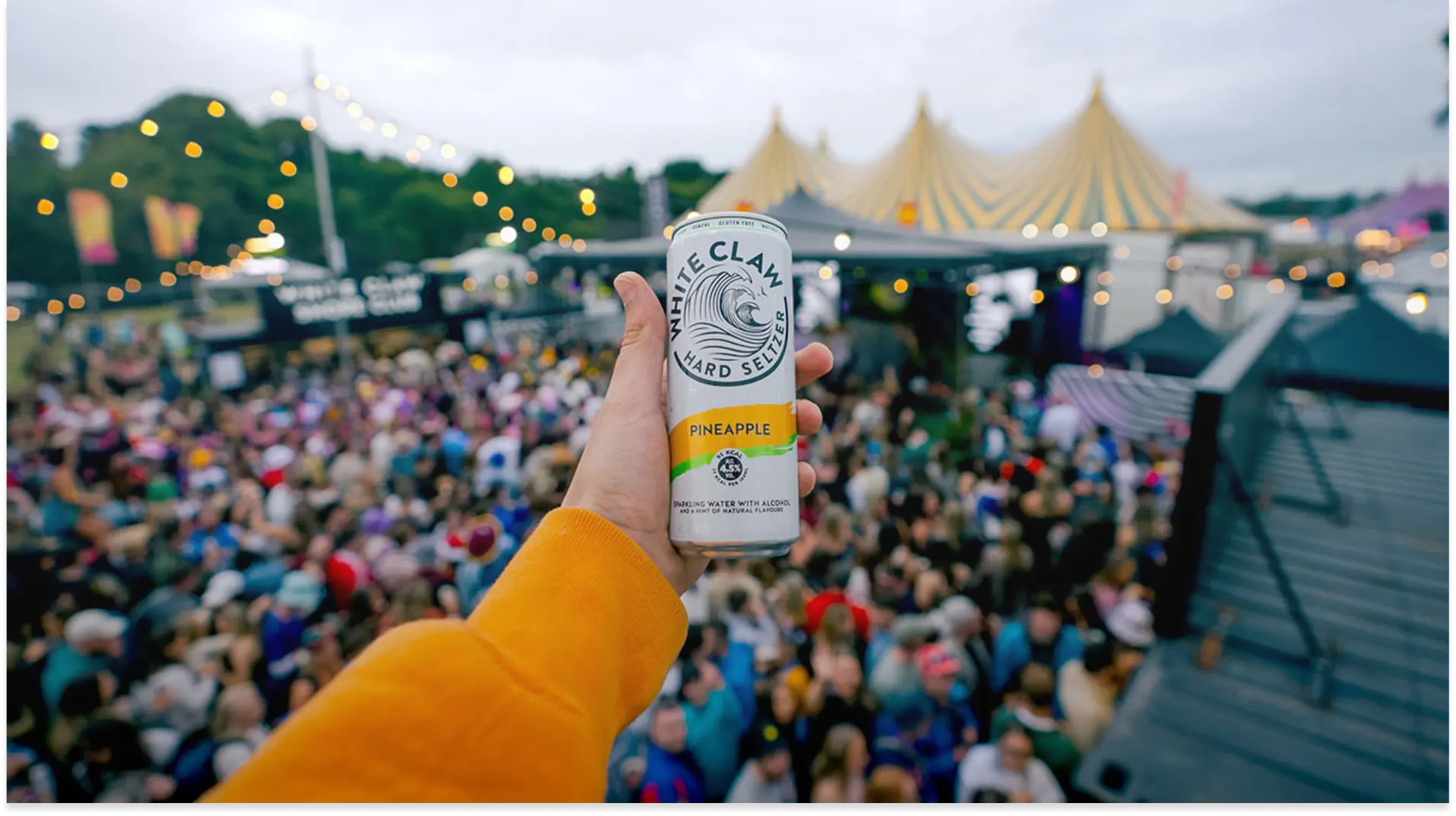 Hand in orange sleeve holding a White Claw Pineapple hard seltzer can at crowded outdoor event.