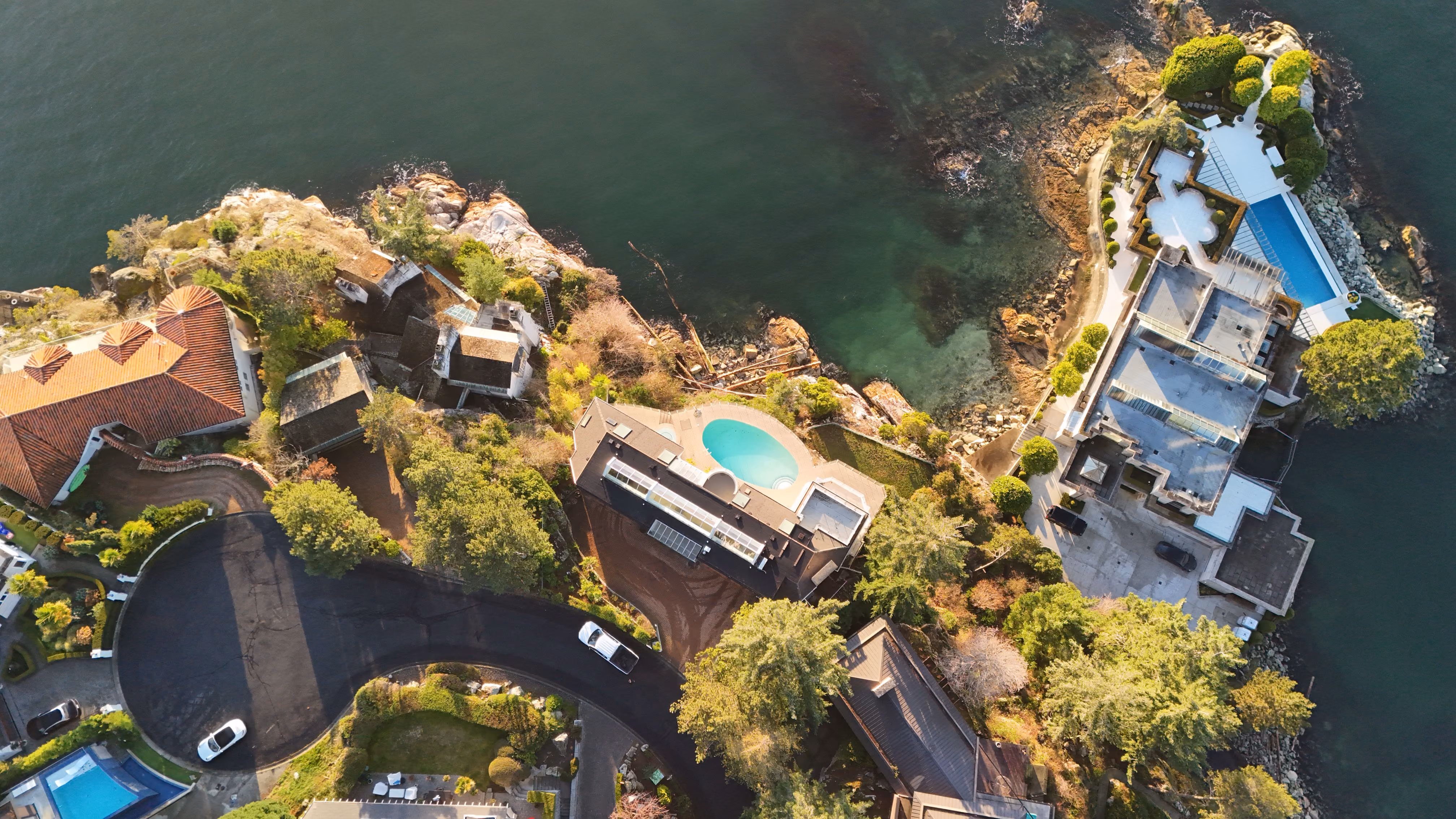 Aerial view of waterfront houses with pools, trees, and rocky shoreline along calm water.