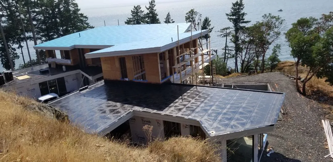 Waterfront home on a rocky bluff with conifers close to the house, torch-on membrane being installed on the lower roof