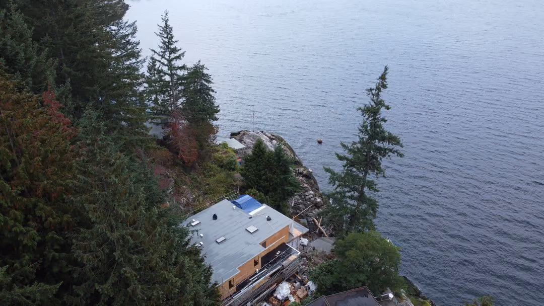 Drone view down a steep treed bluff to a home at the shoreline, tall conifers on all sides