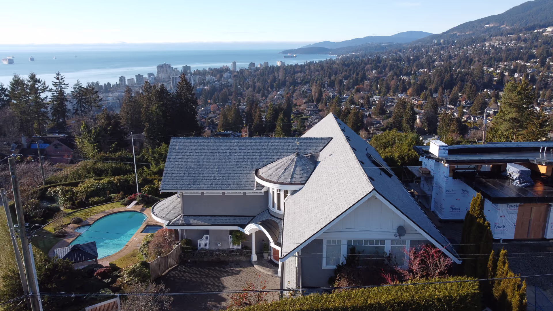 Drone view of a large grey architectural-shingle home with a pool, city and water beyond — Greater Vancouver