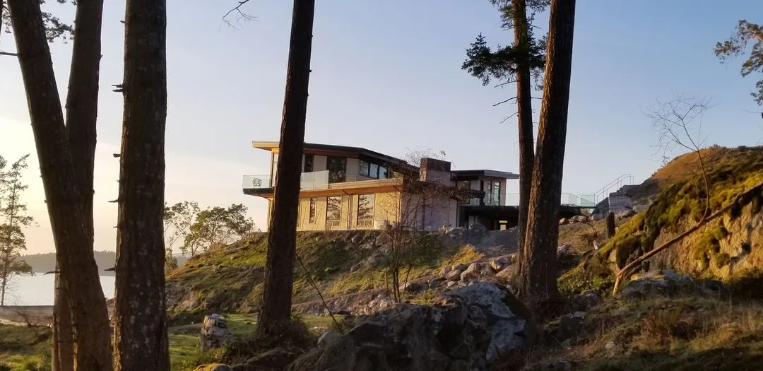 Modern home with a low-slope roof and deep overhang on a mossy rock bluff among pines, water beyond