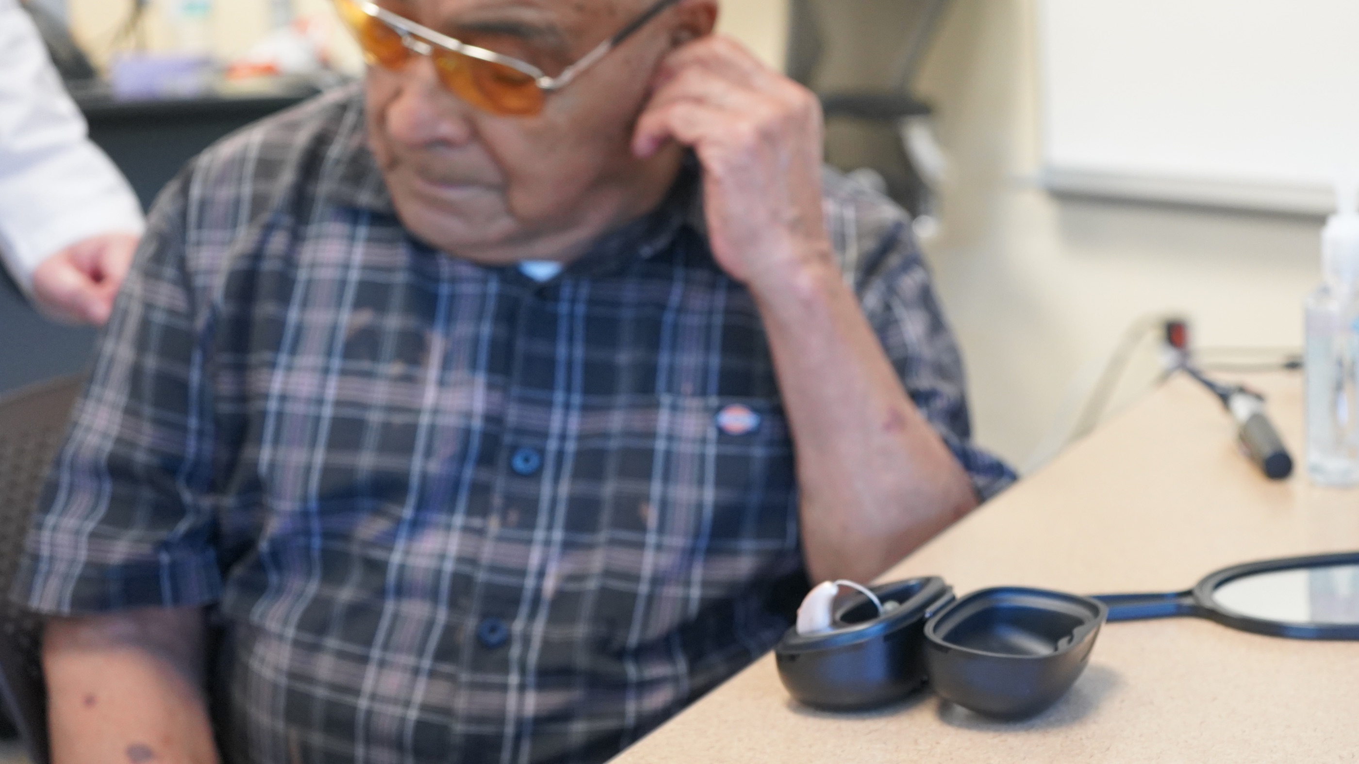 An older man adjusts his newly fitted hearing aid at a clinic desk