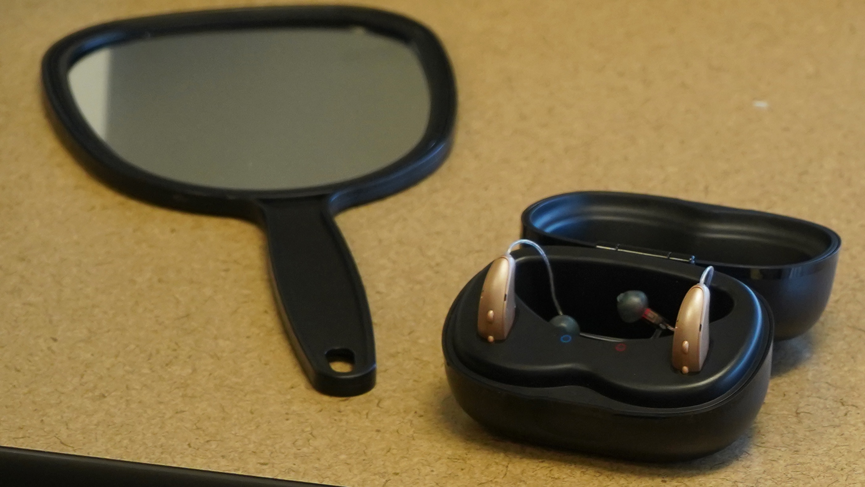 Two hearing aids in an open charging case beside a hand mirror