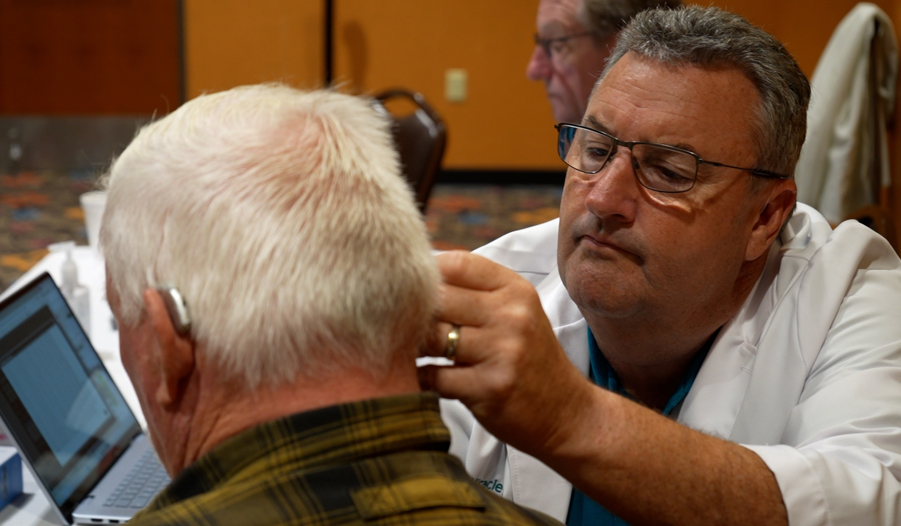 A specialist fits a hearing aid behind an older man's ear beside a laptop